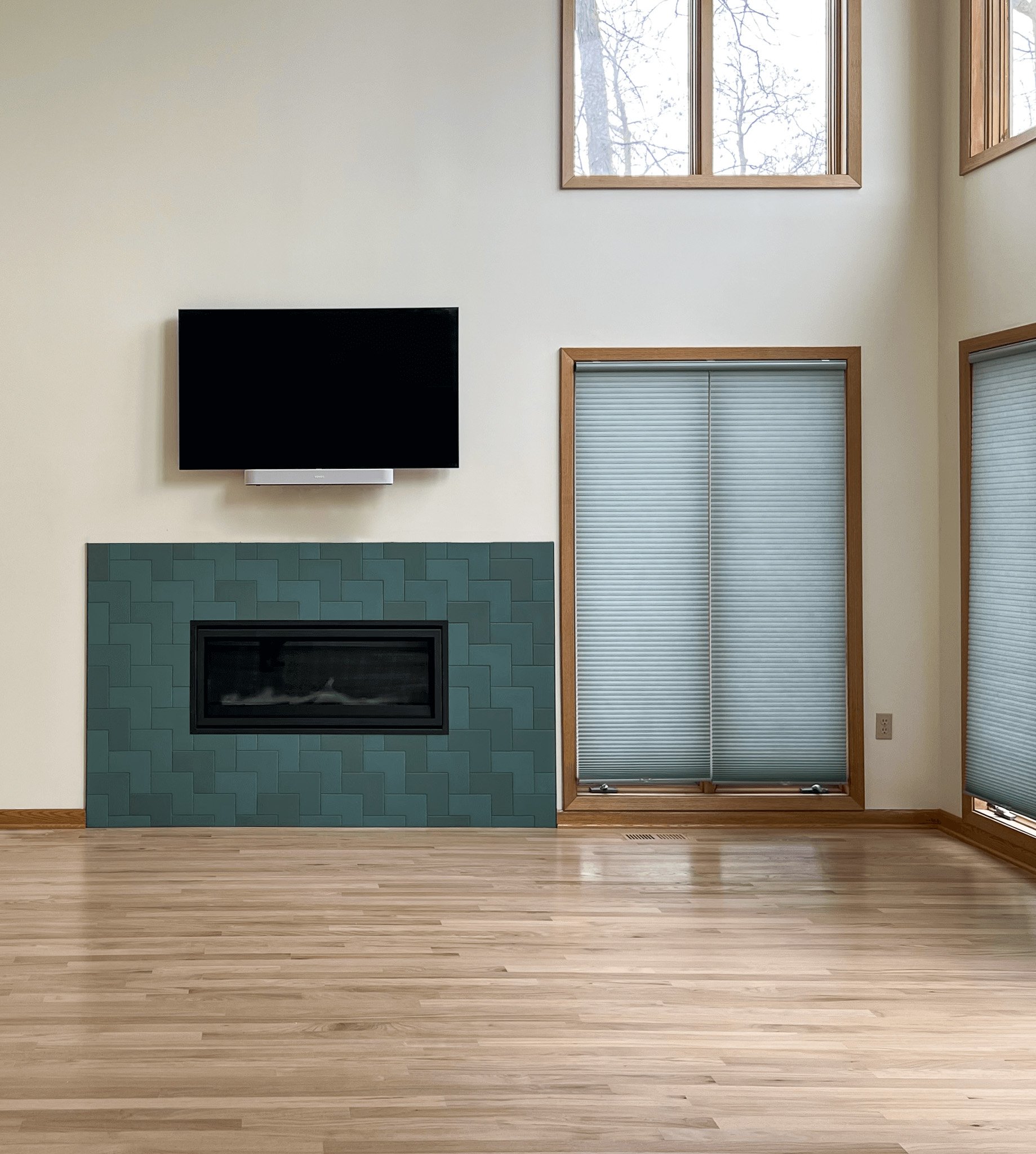 A modern living room featuring a striking fireplace with a monochromatic teal tile surround arranged in a bold geometric pattern. The large format tiles provide texture and visual interest, adding a sleek, contemporary touch to the minimalist design. Natural light pours in from the tall windows, reflecting off the light hardwood floors, creating a bright and airy atmosphere. The mounted flat-screen TV above the fireplace adds to the room's clean, streamlined look, blending modern technology with elegant tile craftsmanship.