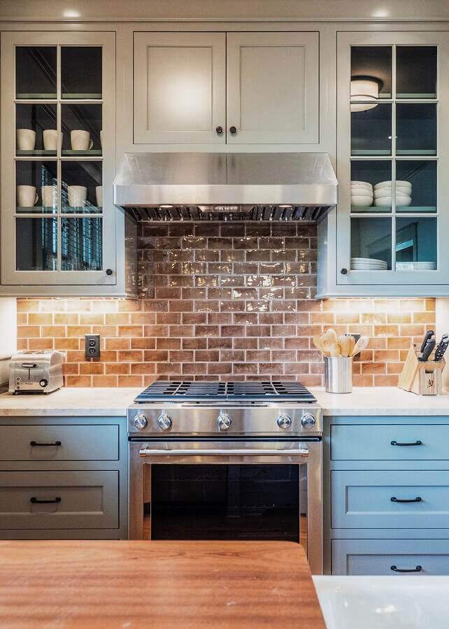 A beautifully designed kitchen featuring a warm, glossy brick-pattern subway tile backsplash in earthy tones. The backsplash pairs seamlessly with soft gray-blue cabinetry and open glass-front upper cabinets, creating a blend of modern and classic design. The stainless steel range and hood complement the neutral color palette, while the marble countertops and under-cabinet lighting enhance the texture and warmth of the space. The overall aesthetic is sleek yet inviting, showcasing the craftsmanship and thoughtful tile selection that elevate the kitchen’s look.