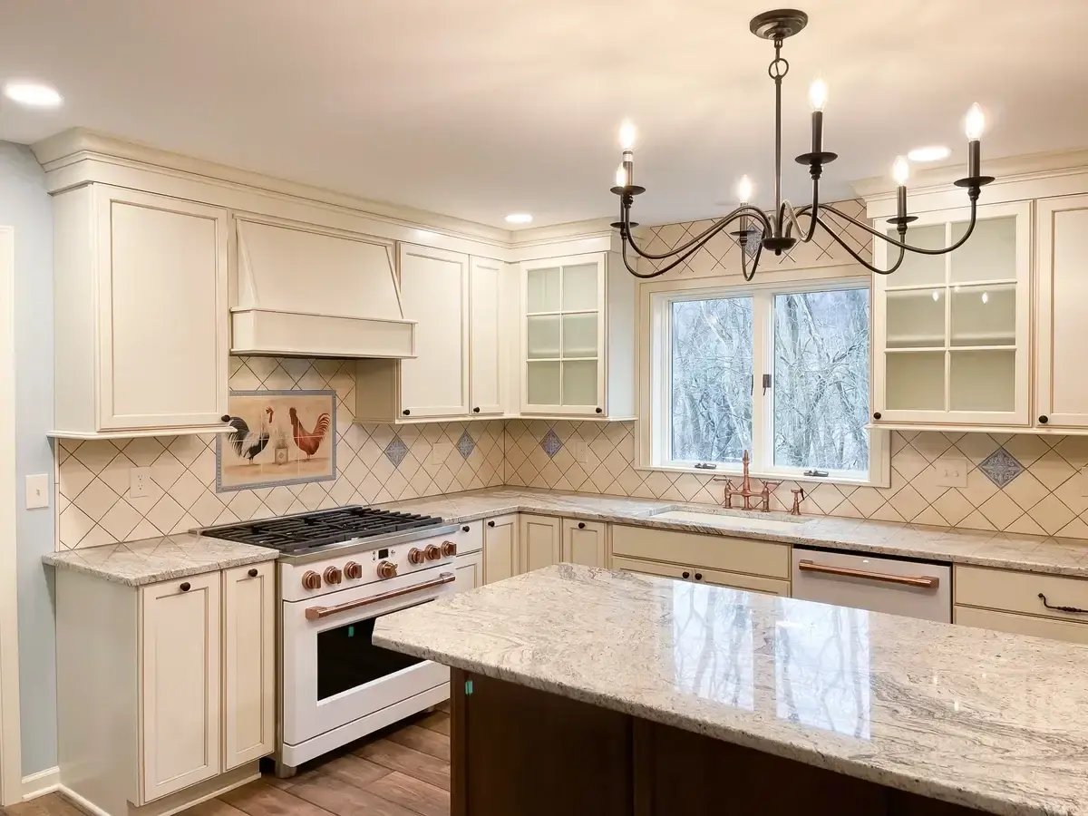 A warm, traditional kitchen featuring cream-colored cabinetry and a custom backsplash with a diamond tile pattern accented by decorative blue tiles. The centerpiece above the stove includes a charming rooster tile mural, adding a touch of farmhouse character to the space. Granite countertops with light veining complement the soft tones of the cabinetry, while a large island provides ample workspace. A classic black wrought-iron chandelier hangs over the kitchen, adding elegance and contrast to the bright, inviting design. Large windows allow natural light to flood the space, highlighting the craftsmanship and details of the kitchen.