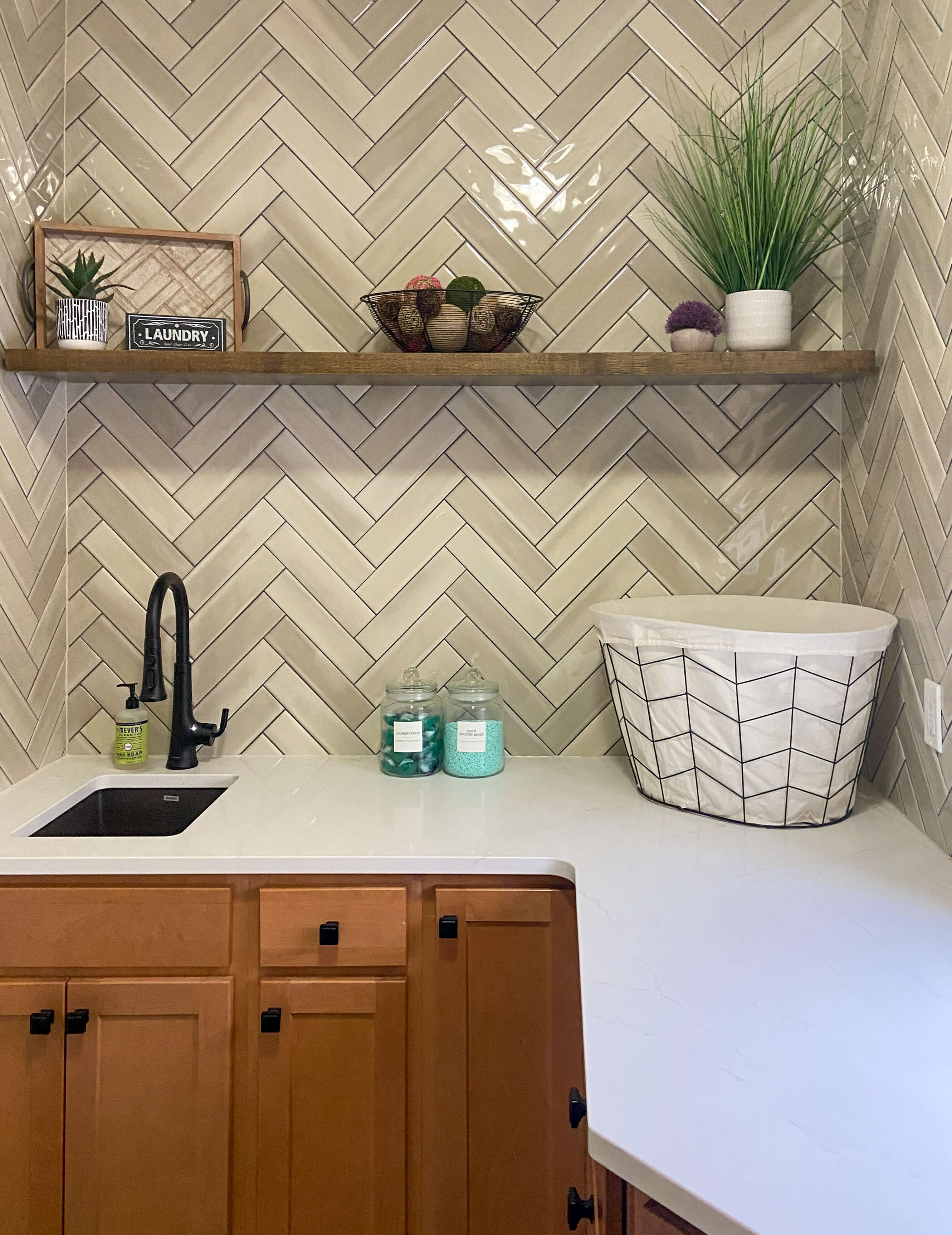 A stylish laundry room featuring a herringbone tile backsplash in soft beige tones, adding texture and visual interest to the space. The wood floating shelf above provides both functionality and warmth, displaying decorative plants and laundry essentials. The rich wood cabinetry contrasts beautifully with the white quartz countertop, which offers ample workspace for laundry tasks. A black matte faucet adds a modern touch, enhancing the overall clean and inviting aesthetic of the room.