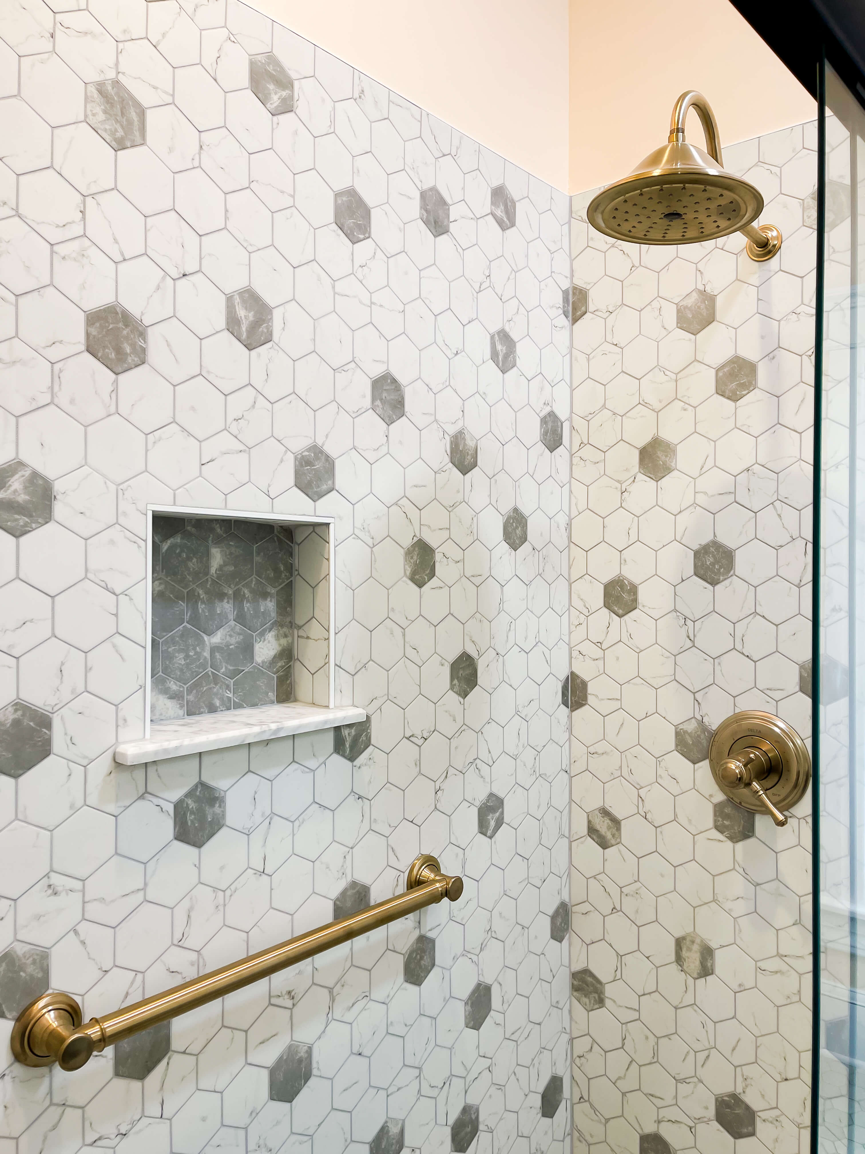 A modern shower featuring a white and gray hexagonal tile pattern with subtle marble veining, creating a sleek and elegant look. The shower includes a recessed niche with coordinating hexagonal tiles for added storage and design continuity. Brass fixtures, including a rain showerhead and a grab bar, add a touch of warmth and luxury, complementing the soft tones of the tile. The overall aesthetic is a blend of contemporary style and classic elegance, offering both functionality and visual appeal.