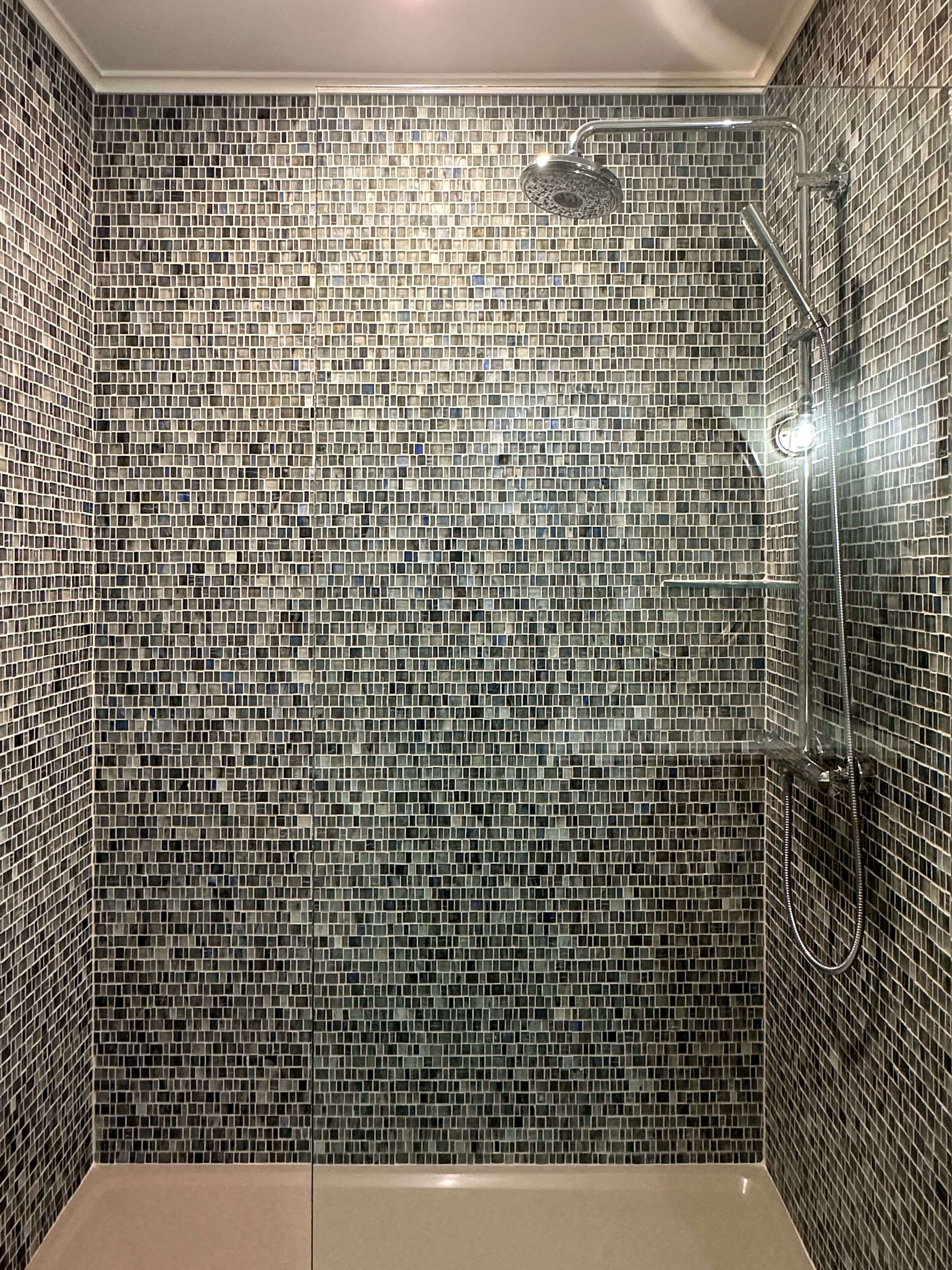 A sleek, modern shower featuring a mosaic tile wall with small, square tiles in varying shades of black, gray, and white, creating a visually striking and textured backdrop. The large rainfall showerhead and handheld attachment, both in polished chrome, add a touch of luxury and complement the reflective qualities of the tiles. The glass partition enhances the open feel of the space, allowing the intricate mosaic tilework to take center stage. The overall design is contemporary and stylish, blending functionality with bold aesthetics.